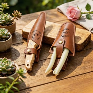 May include: Two gardening tools with tan leather sheaths. One sheath features a circular wreath design with the initials A.F. The other sheath has a floral design with the text Karen's. The tools have light-coloured wooden and cream handles.