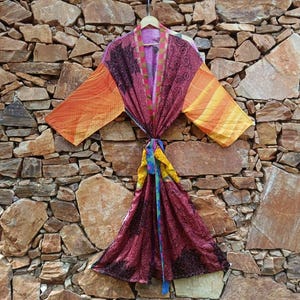May include: A long-sleeved kimono robe featuring a patchwork design with deep burgundy, orange, and yellow panels. The garment displays intricate black floral and swirling patterns throughout, finished with a contrasting patterned trim and a waist tie.