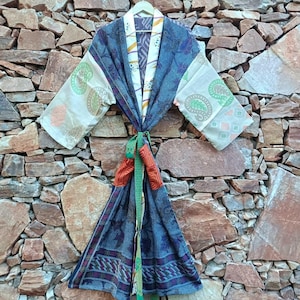以下が含まれることがあります： 青、クリーム色、オレンジ色の様々な布パネルを組み合わせたパッチワークデザインの長袖着物風ローブ。袖には緑色のペイズリー柄があしらわれ、中央には結び紐のベルトがあり、襟元には白いハンガーが掛けられています。