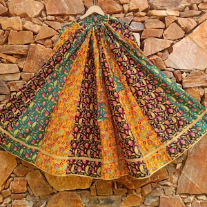 May include: A full-length, flared skirt featuring a vibrant patchwork design with alternating panels of black, yellow, and green fabric. Each panel is decorated with a colorful floral and leaf print, finished with a gold-toned trim along the hem.
