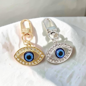 May include: Two eye-shaped charms with lobster claw clasps. One charm features a gold-tone metal frame, while the other has a silver-tone metal frame. Both charms are decorated with clear rhinestones and a central blue, white, and black eye motif.