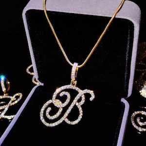 Puede incluir: Un collar de tono dorado con un colgante en forma de letra B cursiva incrustado con piedras claras y brillantes. El colgante cuelga de una cadena dorada a juego, presentado dentro de un joyero forrado de terciopelo gris oscuro.