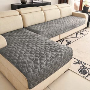 May include: A dark grey quilted sofa cover with a geometric diamond pattern draped over a cream-coloured sectional sofa. The cover features a textured, stitched design that covers the seat cushions and the chaise lounge section of the furniture.