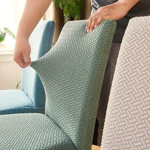 May include: A sage green stretchable chair cover featuring a textured basketweave pattern. The fabric is pulled taut to demonstrate its elasticity and fit over the backrest of a chair. Additional chair covers in blue and white are visible.