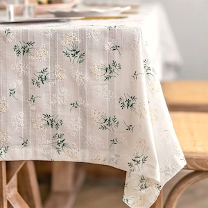 May include: A white tablecloth featuring a vertical striped texture and an embroidered pattern of small green leaves and clusters of tiny white flowers. The fabric drapes over the edge of a table, displaying the repeating botanical design throughout.