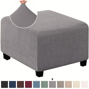 May include: A square ottoman covered in a grey, textured, grid-patterned fabric. The cover features a stretchy, waffle-like weave that conforms to the shape of the ottoman. Four dark, block-style feet support the base of the furniture piece.