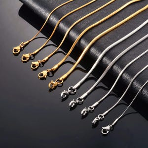 Puede incluir: Una colección de cadenas de serpiente delgadas en tonos dorados y plateados, cada una con un cierre de mosquetón en el extremo. Las cadenas están dispuestas en paralelo, mostrando su textura suave y flexible y su acabado metálico.