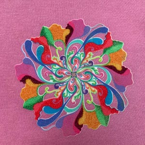 Op de afbeelding: Een roze kledingstuk van stof met een grote, ronde, veelkleurige grafische print. Het ontwerp bestaat uit ingewikkelde, wervelende en bloembladachtige vormen in levendige tinten rood, oranje, geel, groen, blauw en paars met een textuur.