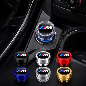 May include: A collection of circular car cigarette lighter plugs featuring the M logo with blue, purple, and red stripes. The set includes various colors such as black, silver, red, blue, clear, and gold, with one plug installed in a car console.