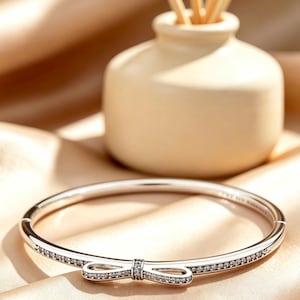 May include: A silver-toned bangle bracelet featuring a central bow-shaped ornament encrusted with small clear stones. The band is also embellished with a row of clear stones along the front, creating a continuous sparkling effect on the metal surface.