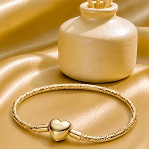 Puede incluir: Una pulsera de cadena de serpiente en tono dorado con un cierre en forma de corazón que tiene la palabra PANDORA grabada en el frente. La pulsera es circular y tiene un acabado metálico pulido, mostrando un diseño de eslabones texturizados.