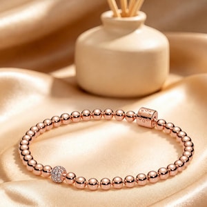 May include: A rose gold-toned beaded bracelet featuring a series of polished spherical beads, a single bead encrusted with small clear stones, and a cylindrical clasp engraved with the word PANDORA, arranged in a circular loop.