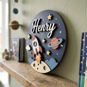 Op de afbeelding: Een ronde houten wandplaat met een donkerblauwe achtergrond en een ruimtethema. De naam Henry is in witte schrijfletters geschreven. Het ontwerp bevat een raket, een planeet met ringen, gele sterren en een maanoppervlak in lichte houttinten.