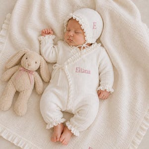 May include: A cream-colored long-sleeve baby romper with ruffled trim, a matching bonnet with the letter E, and a blanket, all featuring pink embroidery of the name Eliana. A beige plush rabbit toy with a pink ribbon sits beside the baby.