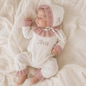 Pode incluir: Um macacão de bebé de manga comprida em cor creme com touca a condizer, com acabamentos em renda rosa seco nos punhos, tornozelos e gola. O macacão tem o nome Ava bordado em fio rosa seco e a touca apresenta a letra A.