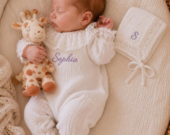 May include: A white long-sleeve romper with a ruffled lace collar and cuffs, featuring the embroidered name Sophia in purple thread. Beside it lies a matching white bonnet with a purple embroidered letter S and a plush giraffe toy.