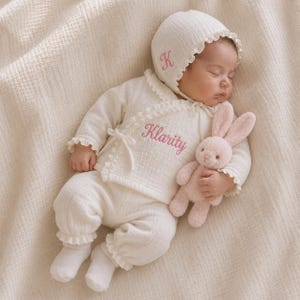 Pode incluir: Um conjunto de blusa transpassada de manga comprida e calças em cor creme com bordas franzidas, bordado com o nome Klarity em rosa. Inclui um gorro a condizer com a letra K rosa e um pequeno coelho de peluche macio em tom rosa.