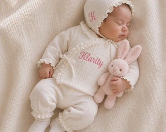 May include: An off-white long-sleeve wrap top and matching pants set with ruffled edges. The top features the embroidered name Klarity in pink, and the matching bonnet displays a pink letter K. A small, soft pink plush rabbit toy is included.