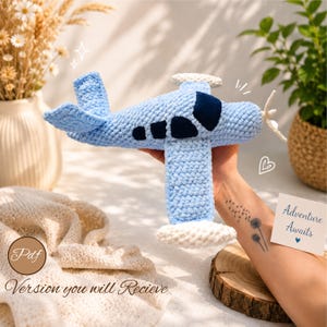 Puede incluir: Un avión de ganchillo azul claro con puntas de ala blancas y una hélice blanca. El avión presenta ventanas y una cabina de color azul oscuro. Una pequeña tarjeta blanca con el texto Adventure Awaits y un icono de corazón azul es visible.