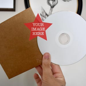 Puede incluir: Una funda cuadrada de cartón marrón para CD junto a un disco circular blanco en blanco. Un gráfico en forma de estrella roja está centrado en la funda con el texto blanco "YOUR IMAGE HERE" impreso dentro. El disco tiene una superficie blanca mate.