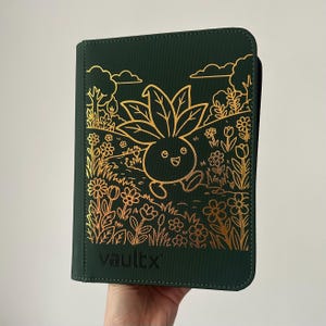 Puede incluir: Un archivador de tarjetas verde oscuro con cremallera que presenta una ilustración de líneas en lámina dorada de una criatura vegetal de dibujos animados caminando por un campo de flores y árboles. El nombre vaultx está grabado en negro.