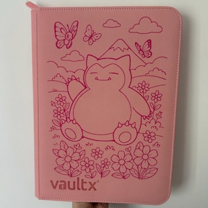 May include: A light pink zipped binder featuring a dark pink line art illustration of a Snorlax character sitting in a field of flowers with butterflies and a mountain in the background. The word vaultx is printed in the bottom left corner.