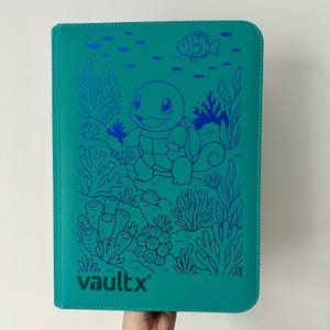 May include: A teal rectangular trading card binder featuring a dark blue line art illustration of a turtle-like creature surrounded by coral, seaweed, and small fish. The brand name vaultx is printed in dark blue at the bottom left corner.