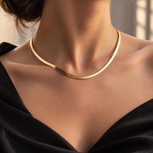 Puede incluir: Un collar de cadena tipo espiga en tono dorado con un diseño de eslabones planos, flexibles y lisos. El collar descansa sobre la clavícula y presenta un acabado metálico pulido que refleja la luz a través de su superficie continua.