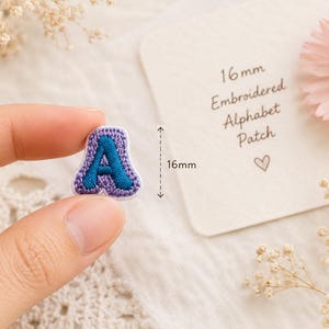 May include: A small embroidered alphabet patch in the shape of the letter A, featuring a dark blue centre and a light purple border. The patch measures 16mm in height as indicated by the text on the adjacent card, which reads 16mm Embroidered Alphabet Patch.