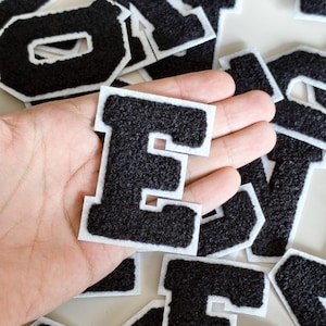 Può includere: Una collezione di toppe a forma di lettera in stile varsity, di colore nero, con una superficie testurizzata e soffice e un bordo bianco. Ogni lettera è tagliata singolarmente e presenta un design a caratteri cubitali. Le toppe sono sparse.
