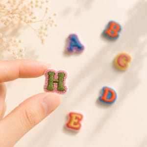 Peut inclure: Un petit écusson brodé en forme de lettre H avec un centre vert et une bordure rose. D'autres écussons brodés en forme de lettres, notamment A, B, C, D et E, sont disposés à proximité, chacun présentant des combinaisons de couleurs distinctes.