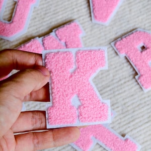 May include: A pink chenille letter patch with a white felt border. The letter K is held in the foreground, while other pink varsity-style letter patches are scattered in the background. The texture of the chenille is fuzzy and raised.
