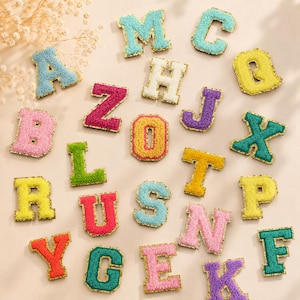 Op de afbeelding: Een verzameling individuele alfabetletters met een getextureerd, pluizig oppervlak in diverse kleuren zoals roze, geel, blauw, groen, oranje en paars, elk omlijnd met een dunne, metallic gouden rand voor decoratieve toepassing.