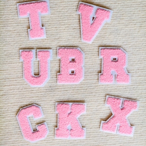 Puede incluir: Una colección de ocho parches de letras individuales con una superficie rosa texturizada y difusa con un borde blanco. Las letras mostradas son T, V, U, B, R, C, K y X, dispuestas en tres filas. Cada letra tiene un diseño de fuente en bloque.