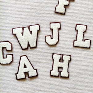 Puede incluir: Un conjunto de seis parches individuales con letras de estilo universitario, con centros de felpilla blanca y bordes de fieltro marrón oscuro. Las letras F, W, J, L, C, A y H están dispuestas de forma dispersa sobre una superficie.