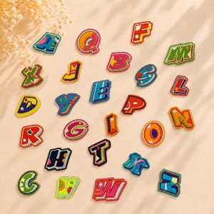 May include: A collection of twenty-six individual embroidered alphabet letter patches. Each patch features a unique, vibrant color scheme and a bold, stylized font design with distinct borders, showcasing the full set of letters from A to Z.