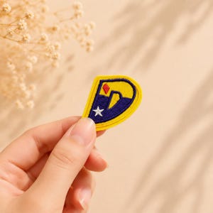 May include: An embroidered patch in the shape of the letter D, featuring a yellow border, a dark blue interior, a small red diamond shape at the top, and a white five-pointed star positioned below the diamond on the blue background.