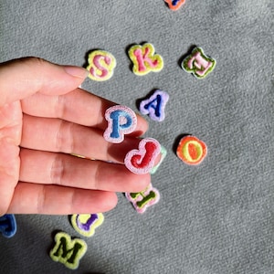 Puede incluir: Una colección de pequeños parches bordados con letras del alfabeto en varios colores brillantes, incluyendo rosa, azul, verde, amarillo y naranja. Cada letra presenta un borde contrastante y está diseñada para su aplicación en telas.