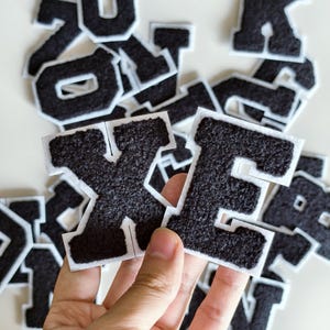 Op de afbeelding: Een verzameling zwarte letterpatches in varsity-stijl met een pluizig, getextureerd oppervlak en een witte rand. De letters liggen op een hoop, waarbij de letters X en E op de voorgrond worden gehouden om het lettertype en materiaal te tonen.