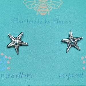 Op de afbeelding: Een paar zilverkleurige zeester-oorstekers met een getextureerd oppervlak en een centraal cirkelvormig detail. De oorbellen zijn bevestigd op een lichtblauwe kaart met de tekst Handmade by Hanna en silver jewellery inspired by nature.