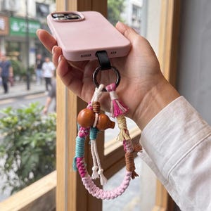 May include: A light pink smartphone case attached to a decorative macrame wrist strap. The strap features braided cords in shades of pink, teal, white, and brown, accented with small tassels and a smooth, polished wooden gourd-shaped charm.
