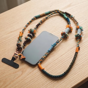May include: A smartphone attached to a long, decorative lanyard made of braided and wrapped multi-colored threads. The lanyard features various textures, including small tassels, wrapped sections in orange, blue, and green, and a copper-toned metal clasp.