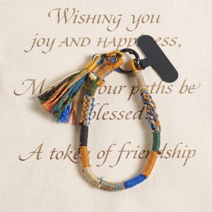 May include: A circular bracelet made of multi-colored thread wrapped around a core, featuring a decorative tassel with blue, green, yellow, and brown strands. A black metal ring connects the bracelet to a small, black, T-shaped plastic tag.