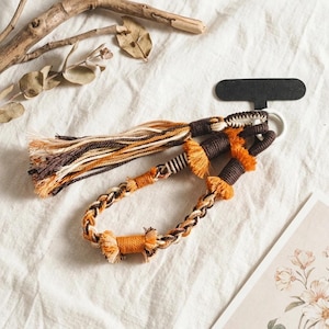 May include: A macrame keychain featuring a braided cord in shades of brown, orange, and cream. The design includes a long tassel, a white metal ring, and a black T-shaped display card. The cord is intricately knotted with wrapped sections.