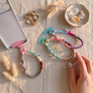 May include: Two phone wrist straps made of multicolored wrapped cord, one attached to a light gray phone case and the other on a blue display card. The straps feature twisted sections in pink, blue, gray, and brown, with small metal rings.