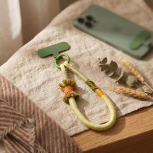 May include: A phone wrist strap featuring a light green cord with wrapped sections in olive, yellow, and orange. It includes a small decorative tassel and a green ring attached to a flat, rectangular green phone anchor tab for device attachment.
