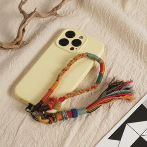 May include: A pale yellow smartphone case with a multi-colored braided cord wrist strap attached to a dark metal ring. The strap features woven patterns in shades of red, orange, blue, and green, ending in a decorative multi-colored fabric tassel.