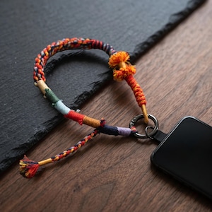 May include: A phone lanyard featuring a braided cord with multi-colored thread wrappings in orange, red, blue, and yellow. The cord includes a decorative orange tassel and a dark metal circular ring attached to a black smartphone corner.