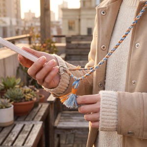 May include: A braided phone lanyard featuring a mix of light blue, white, and orange threads. The strap connects to a metal ring attached to a smartphone and includes a decorative tassel at the base, crafted from matching blue, white, and orange fibers.