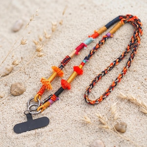 May include: A phone lanyard featuring a braided cord in shades of orange, black, and red. The strap includes decorative wrapped sections in gold, red, and blue, accented with small orange tassels and two silver metal rings attached to a black tab.